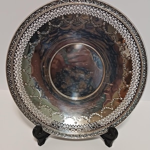 May include: A round silver-toned metal tray featuring a wide, intricate lattice-work border. The inner surface is smooth and reflective, decorated with a delicate embossed garland pattern that follows the circular shape of the tray's rim.