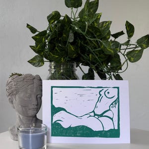 May include: A green and white linocut print of a reclining figure. The print is displayed on a white surface, next to a gray bust, a blue candle in a glass holder, and a jar of green leafy plants.