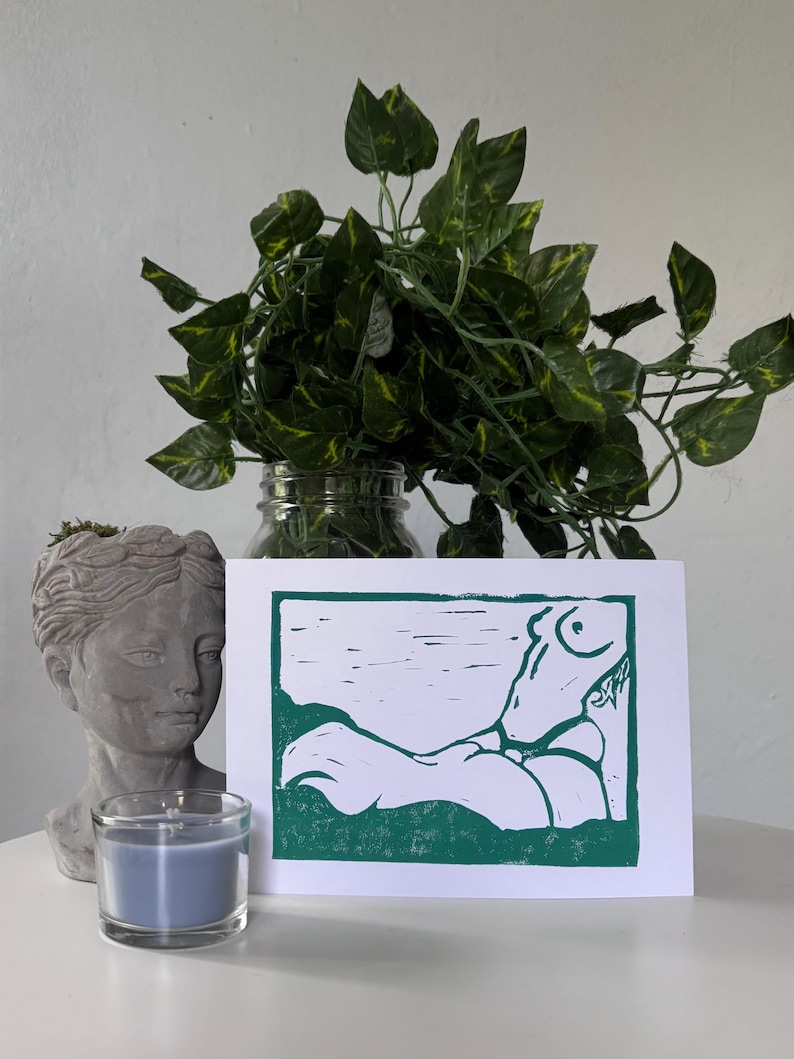 May include: A green and white linocut print of a reclining figure. The print is displayed on a white surface, next to a gray bust, a blue candle in a glass holder, and a jar of green leafy plants.