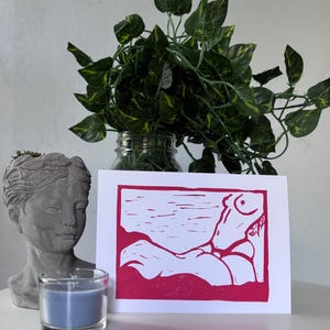 May include: A pink and white art print featuring a stylized figure in a reclining pose. The print is set against a white background and framed by a pink border. A blue candle in a glass holder and a gray bust are in the foreground.