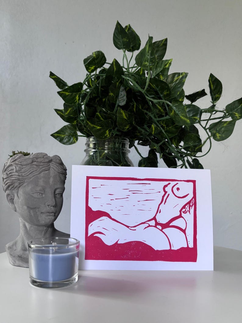 May include: A pink and white art print featuring a stylized figure in a reclining pose. The print is set against a white background and framed by a pink border. A blue candle in a glass holder and a gray bust are in the foreground.
