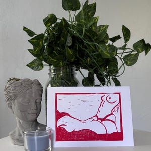 May include: A red and white linocut print of a reclining figure. The artwork is displayed on a white surface, next to a gray bust, a blue candle in a glass holder, and a glass jar with green leafy plants.