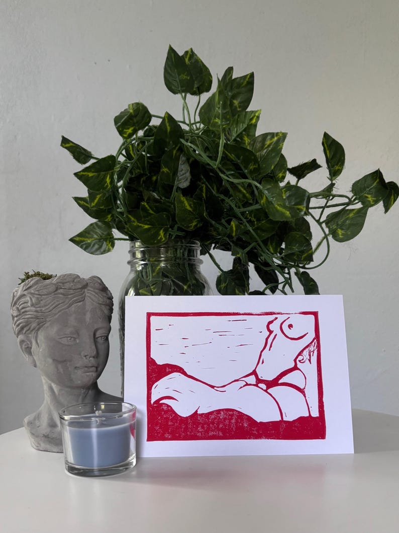 May include: A red and white linocut print of a reclining figure. The artwork is displayed on a white surface, next to a gray bust, a blue candle in a glass holder, and a glass jar with green leafy plants.