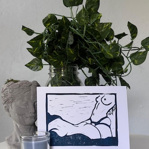 May include: A blue and white linocut print of a reclining figure. The print is displayed with a gray bust, a blue candle, and a glass jar with green leafy vines. The artwork is on a white background.