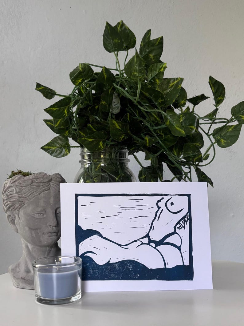 May include: A blue and white linocut print of a reclining figure. The print is displayed with a gray bust, a blue candle, and a glass jar with green leafy vines. The artwork is on a white background.