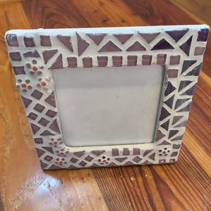 May include: A square photo frame with a mosaic design. The frame is white with purple and red accents. The mosaic pattern includes triangles, squares, and small floral-like details. The frame is designed to hold a photograph.