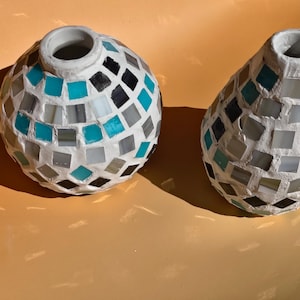 Può includere: Due vasi a mosaico con base bianca e piastrelle di vetro colorate. Uno è rotondo, l'altro a forma di goccia. Le piastrelle sono turchese, grigie, nere e bianche. I vasi hanno una superficie strutturata e un bordo bianco.