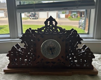 May include: A wooden mantel clock with an ornate, carved design. The clock face is round with black numerals and hands. The clock sits on a rectangular base and is positioned in front of a window.
