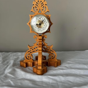 May include: A wooden mantel clock with an ornate, carved design. The clock face is round with Roman numerals and gold-colored hands. The clock is made of light brown wood and has a decorative base and top.