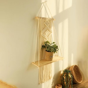 Peut inclure: Étagère murale en macramé crème avec une étagère en bois naturel et une plante en pot. Le motif en macramé présente un motif en losange. L'étagère est suspendue par des cordons crème.