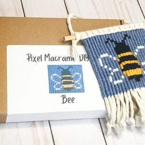 May include: A brown cardboard box with a white label that says "Pixel Macrame DIY Bee". The label also includes a small image of a bee. The box is open and a blue and white macrame wall hanging with a bee design is hanging out of the box.