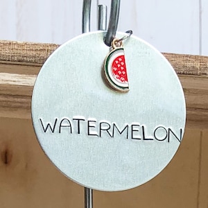 May include: A silver metal tag with the word "WATERMELON" in black lettering. A small watermelon charm hangs from the top of the tag.