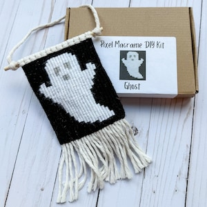May include: A black and white pixel macrame banner featuring a ghost design. The banner has a fringe at the bottom and is suspended from a wooden dowel. A box with a label that reads "Pixel Macrame DIY Kit" and "Ghost" is next to the banner.