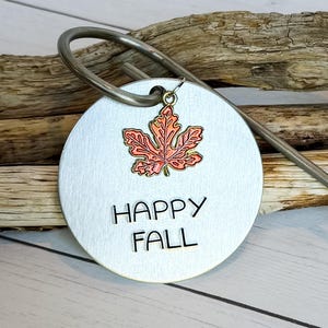 May include: A round, brushed metal garden stake with the words "HAPPY FALL" stamped on it. A small, orange and red leaf charm hangs from the top. The stake has a curved handle.