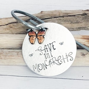 May include: A silver metal charm with the text "Save the Monarchs" and a small orange and black butterfly charm attached. The charm is on a wooden background.