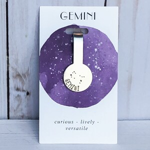 May include: A silver bookmark with a round charm featuring the Gemini constellation and the word "Gemini" engraved on it. The bookmark is on a white card with a purple watercolor background and the words "curious - lively - versatile".