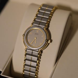 May include: A two-tone wristwatch with a gray face and gold-tone accents. The watch features a silver and gold-tone band and is displayed in a cream-colored box. The watch face has gold-tone hands and markers.