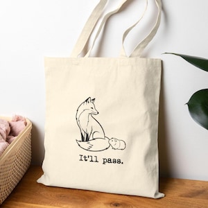 May include: Cream-colored tote bag featuring a black line drawing of a fox and a guinea pig. The text "It'll pass." is printed below the animals. The bag has two long handles and is made of a sturdy fabric.