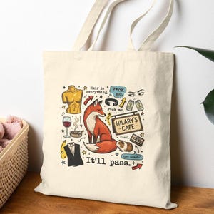 May include: Cream-colored tote bag with a whimsical design featuring a red fox, a guinea pig, and various illustrations. Text includes "Hair is everything," "F*ck me," "Hilary's Cafe," and "It'll pass."