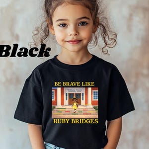 May include: A black short-sleeved t-shirt featuring a graphic print of a school building with the text "BE BRAVE LIKE RUBY BRIDGES" in yellow. The graphic depicts a child walking up steps toward the William Frantz Elementary School entrance.