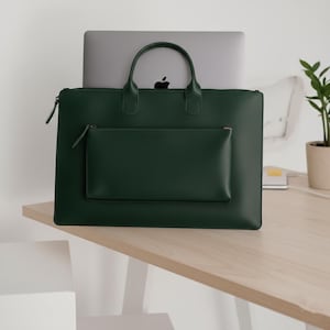 May include: A dark green leather briefcase with a handle and a front pocket. A laptop is partially visible inside the briefcase. The briefcase is on a light-colored wooden surface.