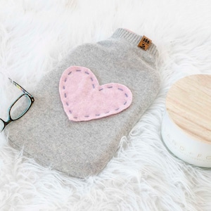 May include: A gray hot water bottle cover with a pink heart applique. The cover is made of a soft material and has a small tag attached. A pair of glasses and a candle are also in the image.