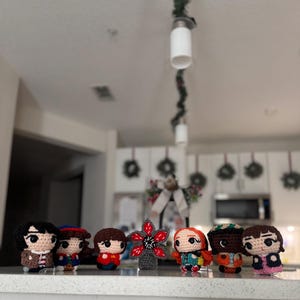 Op de afbeelding: Een verzameling handgemaakte haakfiguren, geïnspireerd op een populaire sciencefiction-serie. De figuren zijn in verschillende kleuren en stijlen, met gedetailleerde gezichtskenmerken en kleding. Een rode en grijze Demogorgon-bloem is ook inbegrepen.