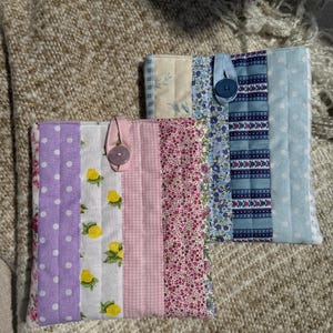 以下が含まれることがあります： ボタン留めの手作り布製ブックカバー2点。1つはラベンダーのドット、レモン柄、ピンクのギンガムチェック柄。もう1つは花柄と青色の生地を使用。どちらも、質感のあるニュートラルな色合いの表面に置かれています。