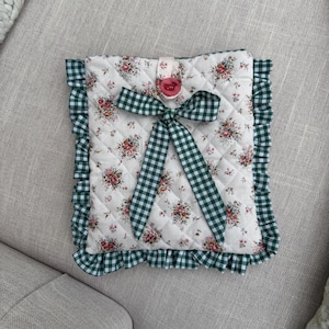 May include: A quilted case with a floral pattern, a green gingham bow, and trim. The case has a white background with a repeating pattern of pink and red roses. A pink button secures the flap.