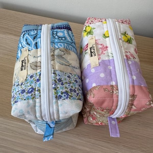 May include: Two quilted pencil cases with white zips. One is blue with floral and paisley patterns, the other is pink, purple, and white with floral and polka dot patterns. Each has a small tag that says "Lauren Studios".