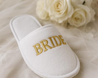 Könnte beinhalten: Weißer Hausschuh mit dem Wort "BRIDE" in goldfarbenem Faden gestickt. Der Hausschuh ist aus einem weichen, plüschigen Material gefertigt. Im Hintergrund weiße Rosen und ein transparenter Stoff.