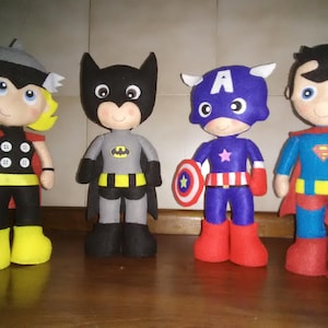 May include: Four handmade superhero dolls: Thor, Batman, Captain America, and Superman. Thor wears a black and yellow outfit with a red cape. Batman is in a gray and black suit. Captain America holds a shield. Superman has a blue and red costume.