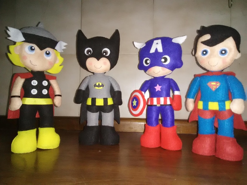 May include: Four handmade superhero dolls: Thor, Batman, Captain America, and Superman. Thor wears a black and yellow outfit with a red cape. Batman is in a gray and black suit. Captain America holds a shield. Superman has a blue and red costume.