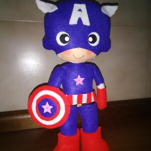 May include: A handmade Captain America doll made of felt. The doll is primarily blue with a white "A" on the head, white wings, and a pink star on the chest. It holds a red, white, and blue shield and wears red boots and gloves.