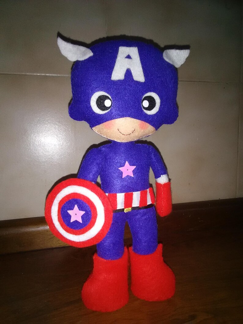 May include: A handmade Captain America doll made of felt. The doll is primarily blue with a white "A" on the head, white wings, and a pink star on the chest. It holds a red, white, and blue shield and wears red boots and gloves.