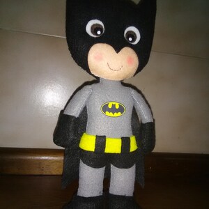 May include: A handmade Batman doll, featuring a black cowl with pointed ears and a smiling face with rosy cheeks. The doll wears a gray suit with a yellow belt and a Batman logo. The doll has a black cape and boots.