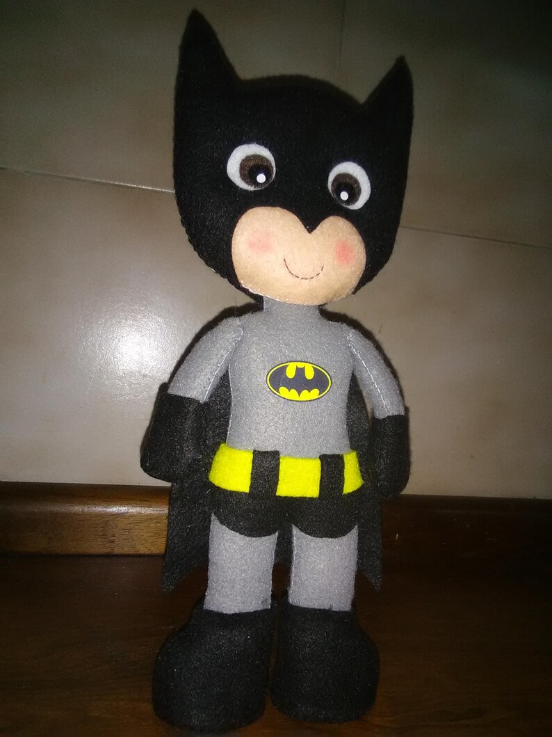 May include: A handmade Batman doll, featuring a black cowl with pointed ears and a smiling face with rosy cheeks. The doll wears a gray suit with a yellow belt and a Batman logo. The doll has a black cape and boots.