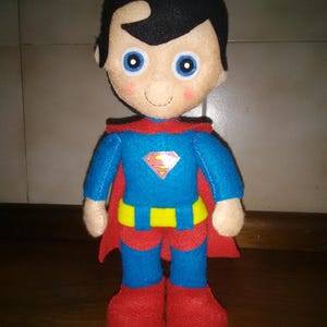 May include: A handmade Superman doll made of felt. The doll has a blue suit with a yellow belt and the Superman logo. It has a red cape, red boots, and black hair. The doll's face has blue eyes and a smiling expression.