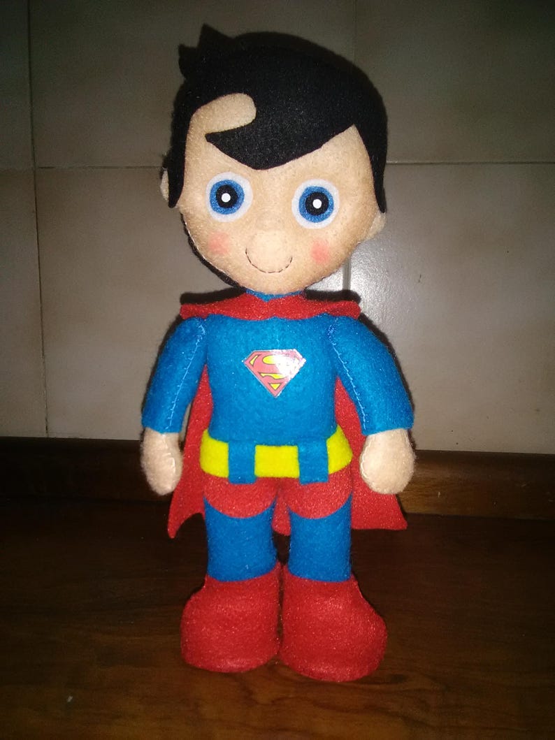 May include: A handmade Superman doll made of felt. The doll has a blue suit with a yellow belt and the Superman logo. It has a red cape, red boots, and black hair. The doll's face has blue eyes and a smiling expression.