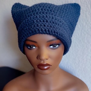 May include: A navy blue crochet hat with pointed cat ears. The hat is made of a textured yarn and is displayed on a mannequin head. The hat is a unique accessory, perfect for adding a touch of whimsy to any outfit.