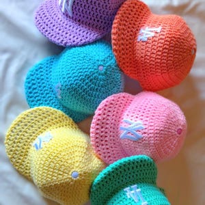 May include: A collection of colorful, crocheted baseball caps in shades of purple, orange, blue, yellow, pink, and green. Each cap features a white embroidered logo on the front and a matching button on top. The caps are arranged in a casual, overlapping pattern.