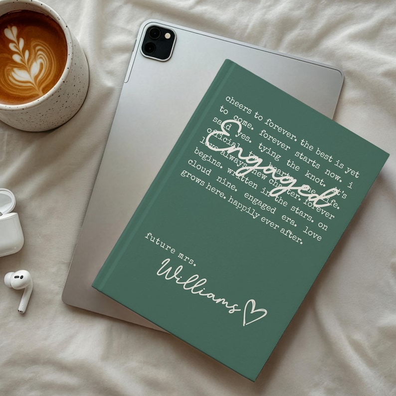 Puede incluir: Un diario verde con la palabra "Engaged" y otro texto en blanco, apoyado en una tableta plateada. El diario tambi&eacute;n tiene las palabras "future mrs. Williams" y un s&iacute;mbolo de coraz&oacute;n. Una taza de caf&eacute; y un par de auriculares blancos tambi&eacute;n est&aacute;n en la imagen.