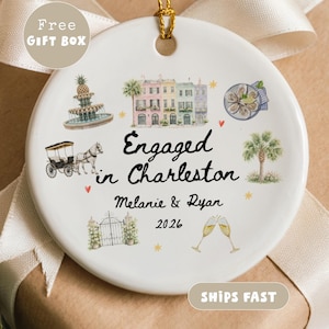 May include: A round white ceramic ornament featuring watercolor illustrations of a fountain, historic buildings, a horse-drawn carriage, a palm tree, oysters, and champagne flutes. The text reads Engaged in Charleston, Melanie & Ryan, 2026.