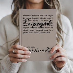 Puede incluir: Un diario beige con la palabra "Engaged" en escritura cursiva. El diario tiene texto que dice "cheers to forever...happily ever after." Debajo del texto est&aacute; "future mrs. Williams" con un s&iacute;mbolo de coraz&oacute;n.