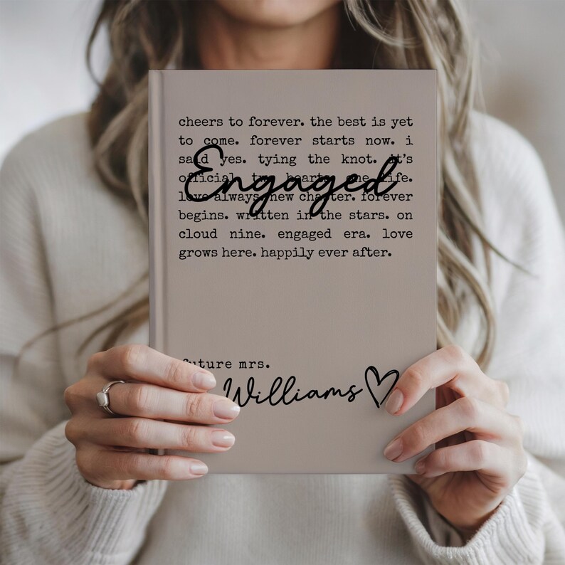 Puede incluir: Un diario beige con la palabra "Engaged" en escritura cursiva. El diario tiene texto que dice "cheers to forever...happily ever after." Debajo del texto est&aacute; "future mrs. Williams" con un s&iacute;mbolo de coraz&oacute;n.