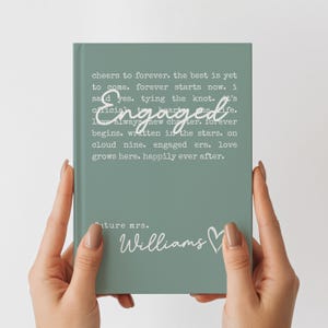 Puede incluir: Un cuaderno verde salvia con la palabra "Engaged" en escritura cursiva. El cuaderno tiene el texto "cheers to forever...happily ever after" y "future mrs. Williams" en blanco. El cuaderno est&aacute; sostenido por dos manos.