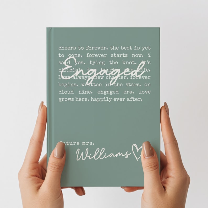 Puede incluir: Un cuaderno verde salvia con la palabra "Engaged" en escritura cursiva. El cuaderno tiene el texto "cheers to forever...happily ever after" y "future mrs. Williams" en blanco. El cuaderno est&aacute; sostenido por dos manos.