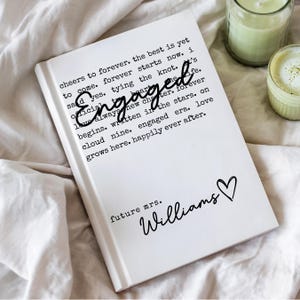 Puede incluir: Un diario de tapa dura blanco con la palabra "Engaged" en cursiva y texto sobre el amor y el matrimonio. El diario incluye las palabras "future mrs. Williams" y un s&iacute;mbolo de coraz&oacute;n. El diario est&aacute; sobre una superficie blanca.