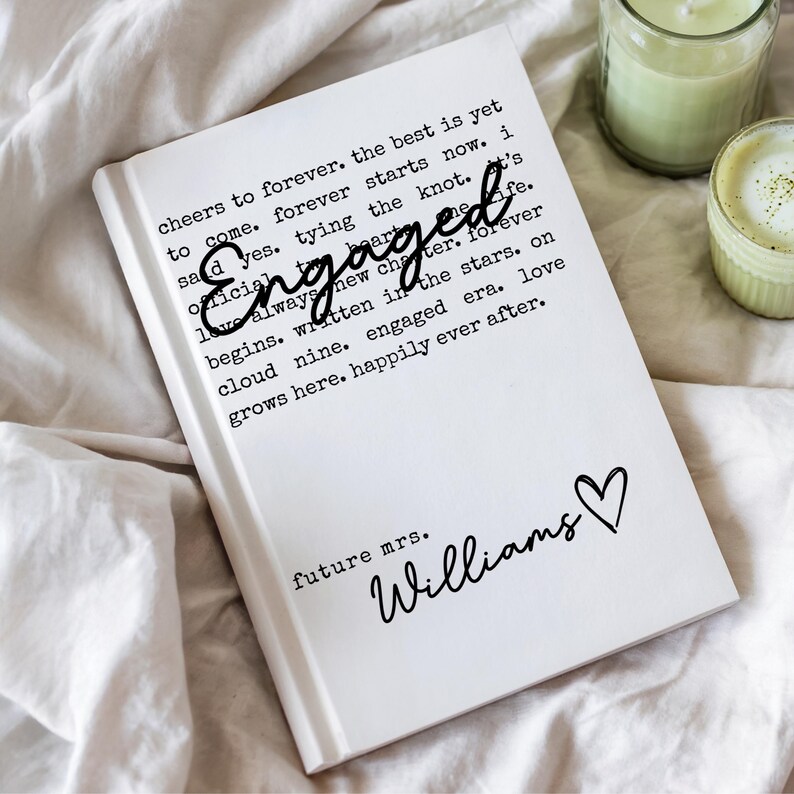 Puede incluir: Un diario de tapa dura blanco con la palabra "Engaged" en cursiva y texto sobre el amor y el matrimonio. El diario incluye las palabras "future mrs. Williams" y un s&iacute;mbolo de coraz&oacute;n. El diario est&aacute; sobre una superficie blanca.