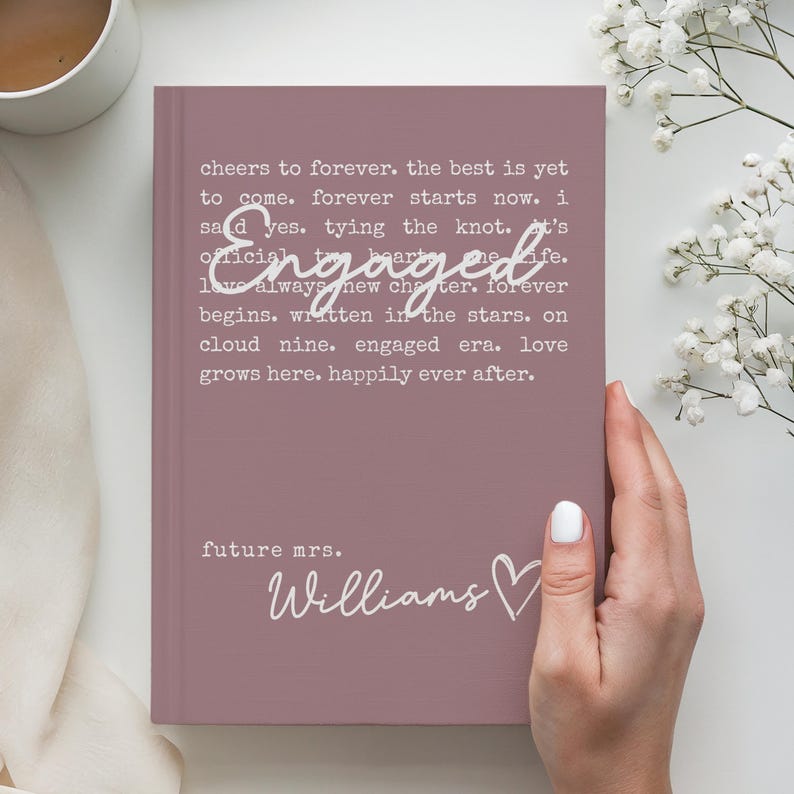 Puede incluir: Un diario de color malva con la palabra "Engaged" en una fuente de escritura. El diario tiene texto que dice "cheers to forever" y "future mrs. Williams" con un s&iacute;mbolo de coraz&oacute;n. El diario es sostenido por una persona.